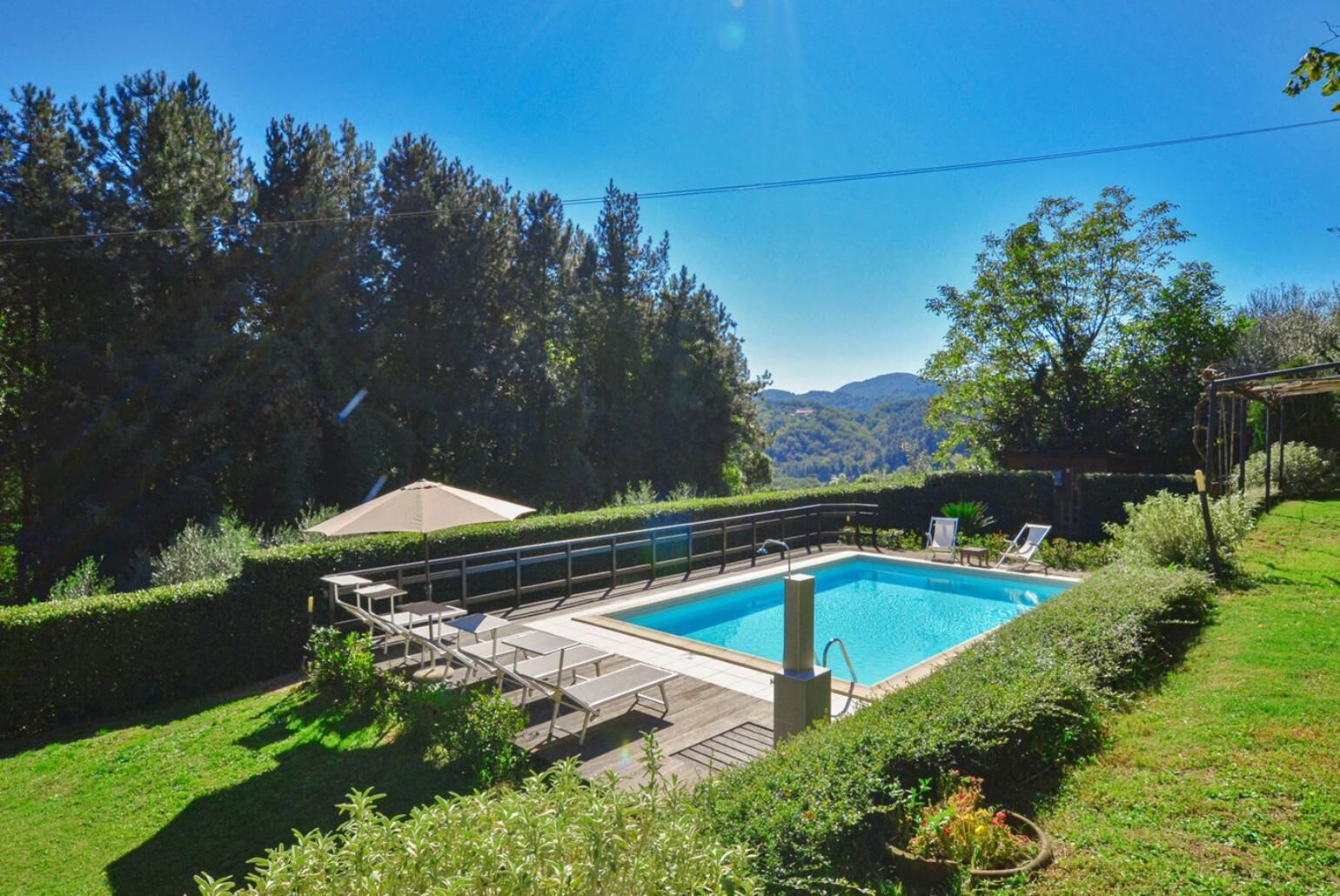 A rectangular swimming pool with a wooden deck and lounge chairs, surrounded by well-maintained hedges and a view of a mountainous landscape.