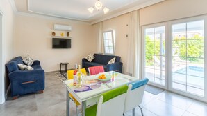 Un salon moderne comprenant un canapé, une table à manger avec des chaises, un téléviseur fixé au mur et une grande porte-fenêtre coulissante donnant sur un espace extérieur.