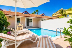 Une piscine entourée de chaises longues et d’un parasol, le tout bordé par un bâtiment doté d’un toit en tuiles et d’une porte.