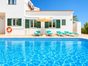 A swimming pool with clear blue water, surrounded by a white building with green shutters, and several lounge chairs.