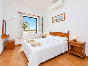 A bedroom with a bed, a nightstand, a bedside table, a window with curtains, a wall-mounted air conditioner, and a framed picture.