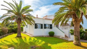 Une villa blanche, dotée d’un toit en tuiles, entourée d’une pelouse bien entretenue et de palmiers.