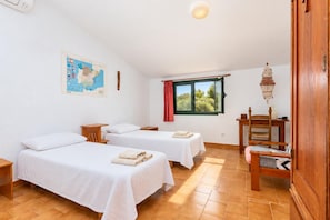 A room with two beds, a wooden chair, a table, a map on the wall, and a window with a view of greenery.