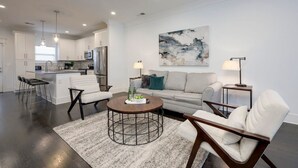 Uma sala de estar moderna com um sofá, poltronas, uma mesa de centro e uma cozinha ao fundo.