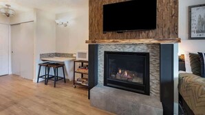 Ein Wohnzimmer mit Kamin, einem flachbildfernseher darüber und einer Küchenzeile mit Arbeitsfläche und Hockern.