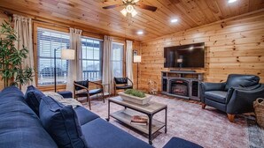A cozy cabin interior with a wooden ceiling, a fireplace, a television, a sofa, and two armchairs.