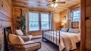 A wooden cabin bedroom with a bed, two chairs, a ceiling fan, and large windows.