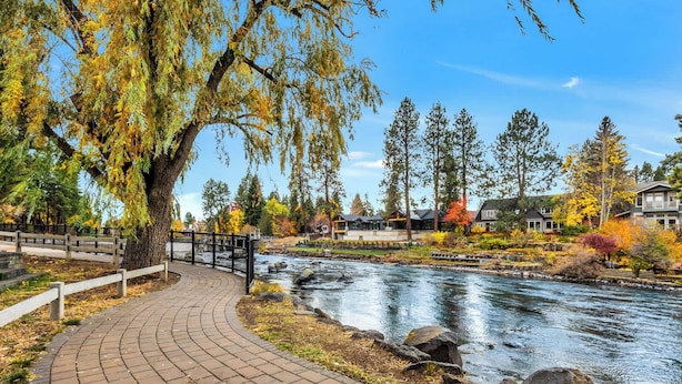 Un paseo junto al río con un puente, rodeado de follaje otoñal y edificios residenciales.