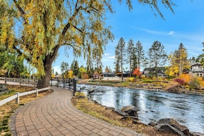 A riverside walkway with a bridge, surrounded by autumn foliage and residential buildings.