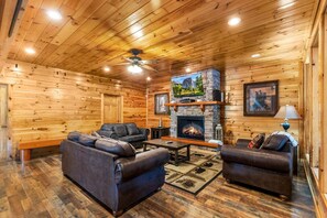 A log cabin interior with a fireplace, leather sofas, a wooden floor, and a ceiling fan. - Splash Mansion - Indoor Pool and Theater (Sevierville)