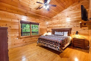 A wooden cabin room with a bed, a nightstand, a ceiling fan, and a window with a view of trees. - Splash Mansion - Indoor Pool and Theater (Sevierville)
