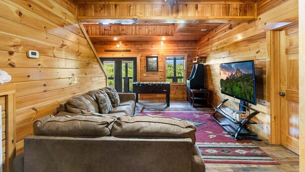 A wooden cabin interior with a sofa, a foosball table, a television, and a ceiling fan. - Splash Mansion - Indoor Pool and Theater (Sevierville)
