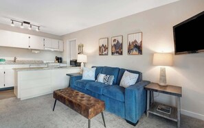 Ein modernes Wohnzimmer mit einem blauen Sofa, einem braunen Hocker, einem Fernseher und Bildern an der Wand.