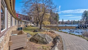 A riverside walkway with a stone bench, a large tree, and a building with large windows.