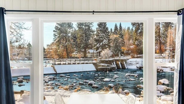 A room with a large window overlooking a river, surrounded by trees and a bridge.