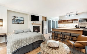 A modern bedroom with a bed, a fireplace, and a kitchen area with bar stools.