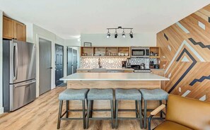 Una cocina moderna con una isla central, taburetes de bar, electrodomésticos de acero inoxidable y un revestimiento de pared con estampado geométrico.