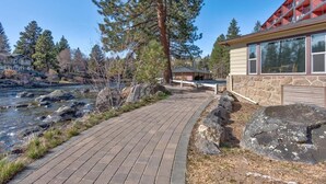 A paved walkway beside a river, a stone building with large windows, and a tall pine tree.