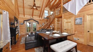 A wooden cabin interior with a dining area, black leather seating, a ceiling fan, and a staircase leading to an upper level.