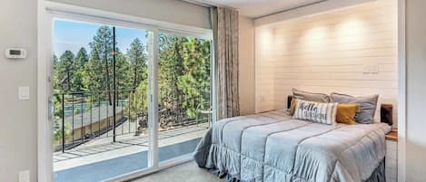 Un dormitorio con un amplio ventanal corredera que da a un balcón, una cama con cubrecama gris y una lámpara fijada a la pared.