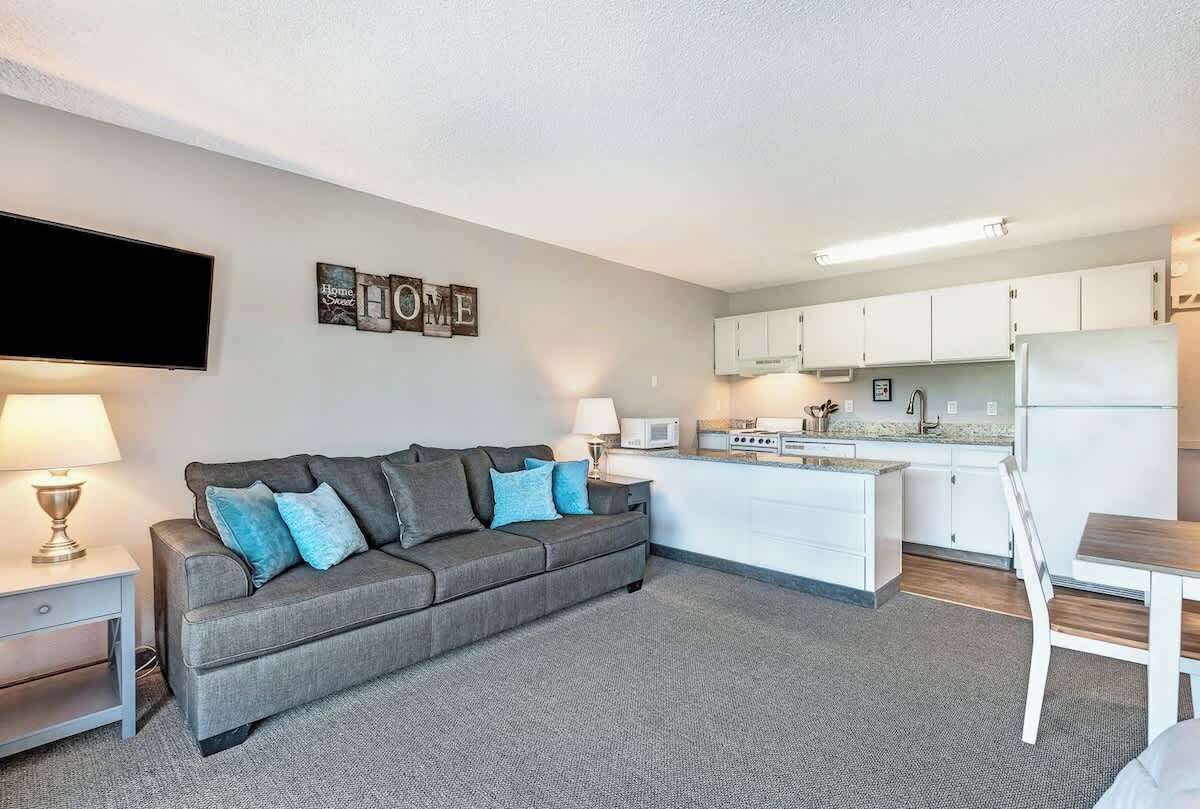 A modern living room with a grey sofa, a flat-screen TV, and a kitchen area with white cabinets and appliances.