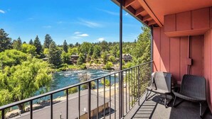 A balcony with a view of a river and forest, two wicker chairs, and a red building.