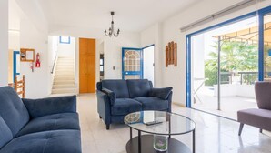 A modern living room with a blue sofa set, a glass coffee table, and a view of a balcony with outdoor furniture. - Villa Charoula Pente (Polis)