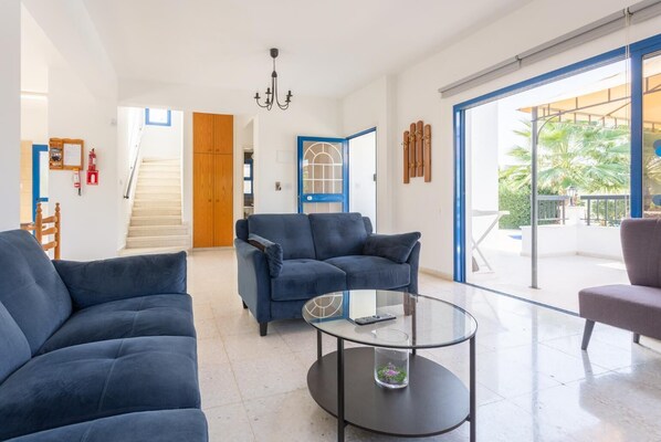 Ein modernes Wohnzimmer mit einer blauen Sofagarnitur, einem Glastisch und Blick auf einen Balkon mit Gartenmöbeln.