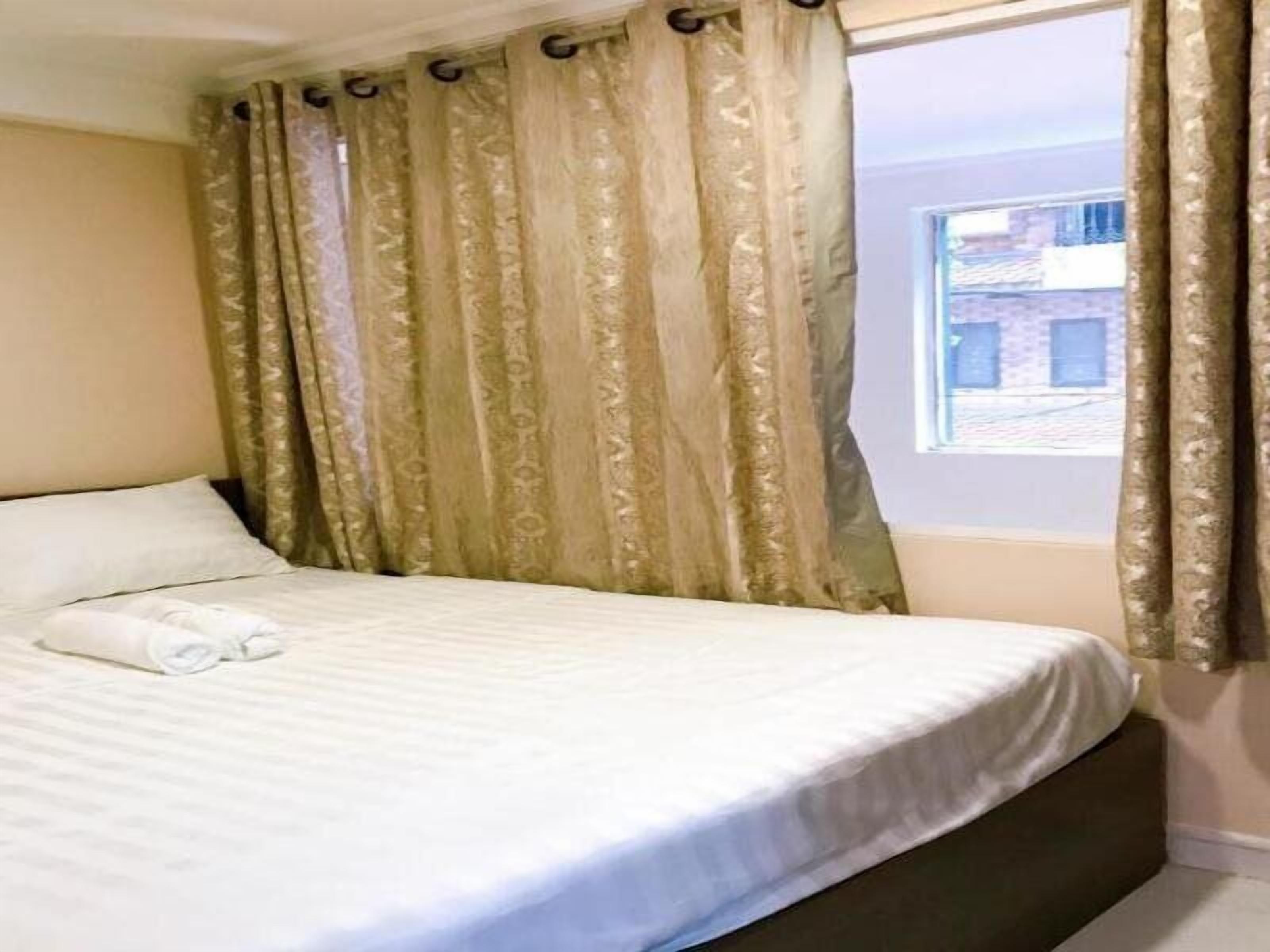 A neatly made bed with a white comforter and a rolled towel, a window with a view of buildings, and a curtain with a floral pattern.
