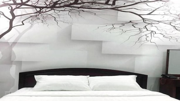 Un lit avec une literie blanche et une tête de lit sombre, une table de chevet avec un miroir et un mur décoré d’un motif représentant une branche d’arbre.