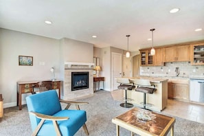 A living room with a fireplace, a blue chair, a coffee table, and a kitchen area with wooden cabinets and a countertop.