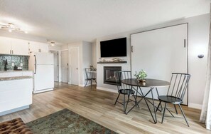 A modern kitchen with white cabinets, a dining area with a round table and chairs, a fireplace, and a flat-screen TV mounted above it.