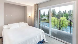 Habitación de hotel con una cama grande, puerta corrediza de vidrio a un balcón y vistas de una zona ajardinada con árboles y un edificio.