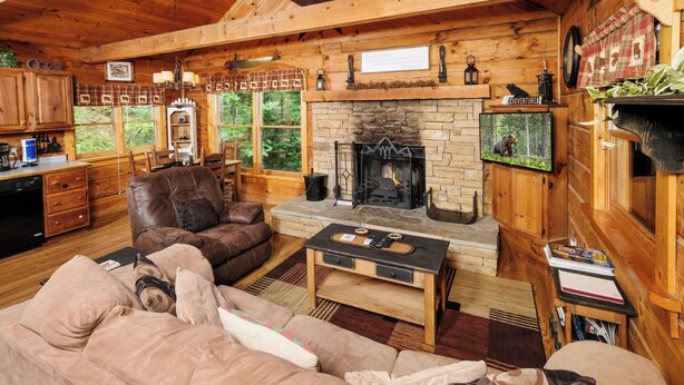 A cozy cabin interior with a stone fireplace, wooden furniture, and a television mounted on the wall.
