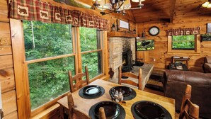 A cozy cabin interior with a dining area, fireplace, and large windows overlooking a forest.