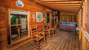 A wooden cabin with a wooden deck, rocking chairs, a hot tub, and a welcome sign.