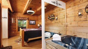 A wooden cabin interior with a bed, a bathroom with a stone wall, and a window with a view of trees.