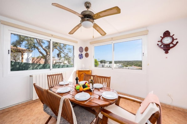 Uma sala de jantar com uma mesa de madeira posta para seis pessoas, um ventilador de teto e vista para a paisagem através de grandes janelas.