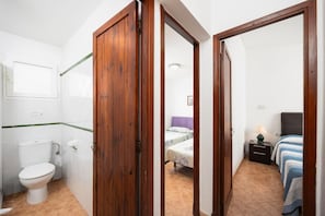 Een kamer met een houten deur, een toilet, een bed met gestreept beddengoed en een nachtkastje met een lamp.