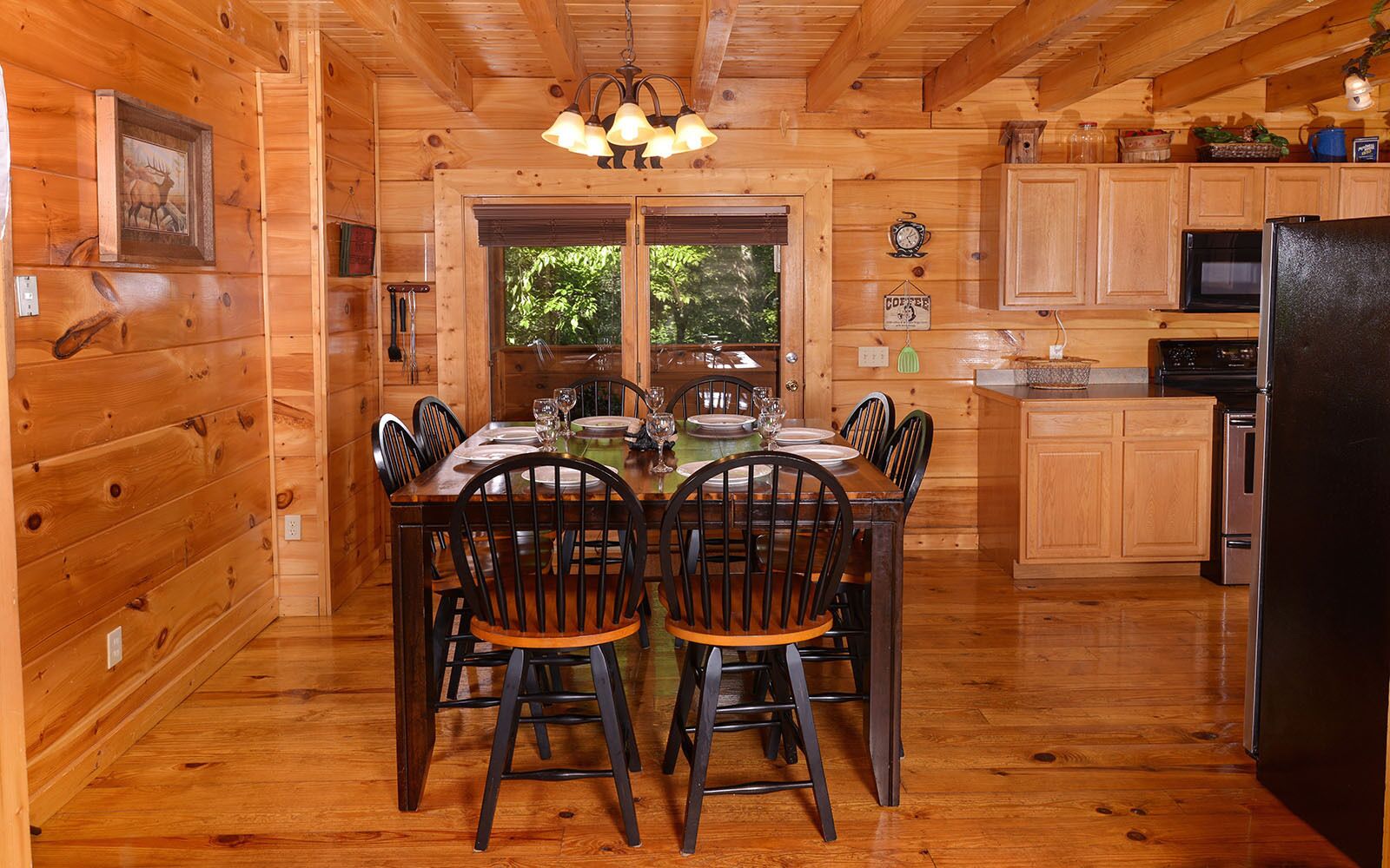 Ein Holzblockhaus-Interieur mit einem Esstisch, gedeckt für vier Personen, Küchenschränken, einem Kühlschrank und einem Fenster mit Blick auf grüne Vegetation.
