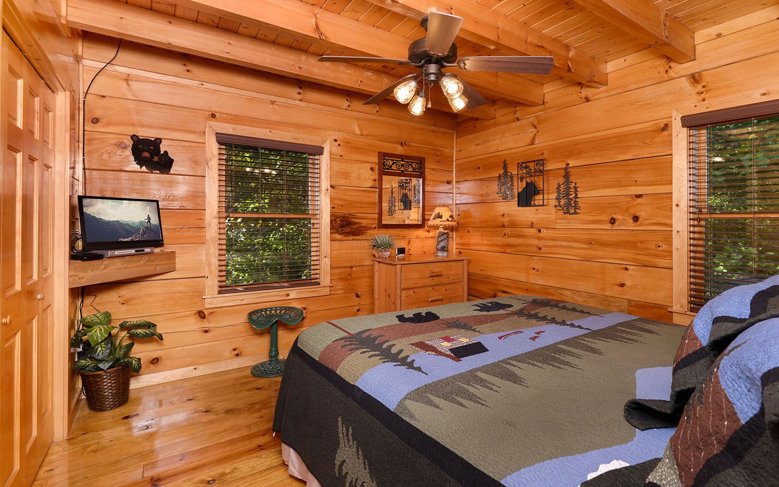 Une chambre confortable dans un chalet, avec un lit, un téléviseur posé sur une étagère, un ventilateur de plafond et une fenêtre avec des stores.