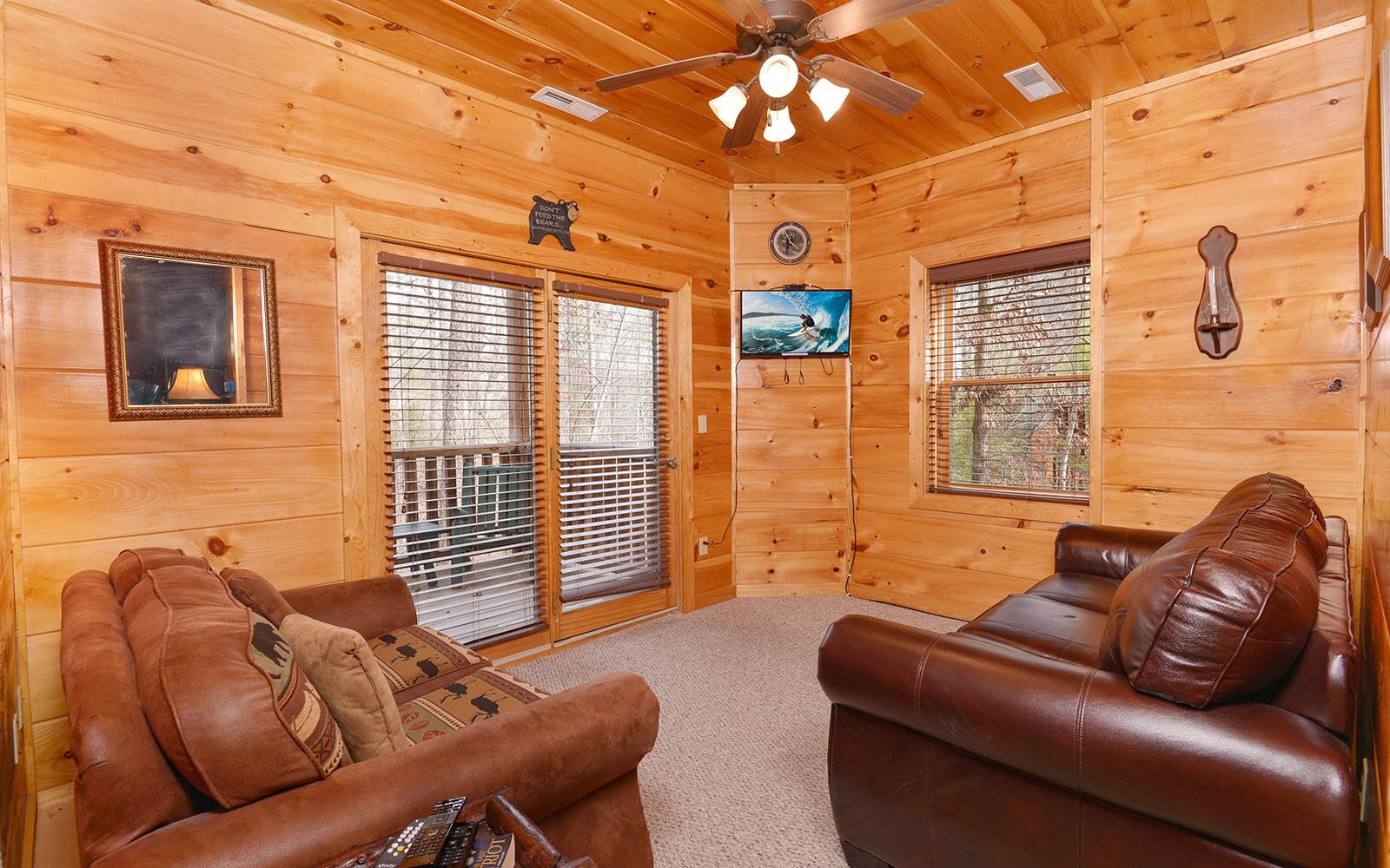 A cozy cabin interior with two leather sofas, a ceiling fan, wooden walls, and large windows with blinds.
