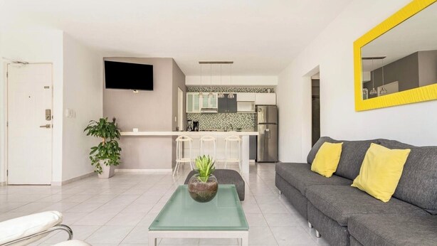 Ein modernes Wohnzimmer mit einem grauen Sofa, gelben Kissen, einem Glastisch und einer Topfpflanze.