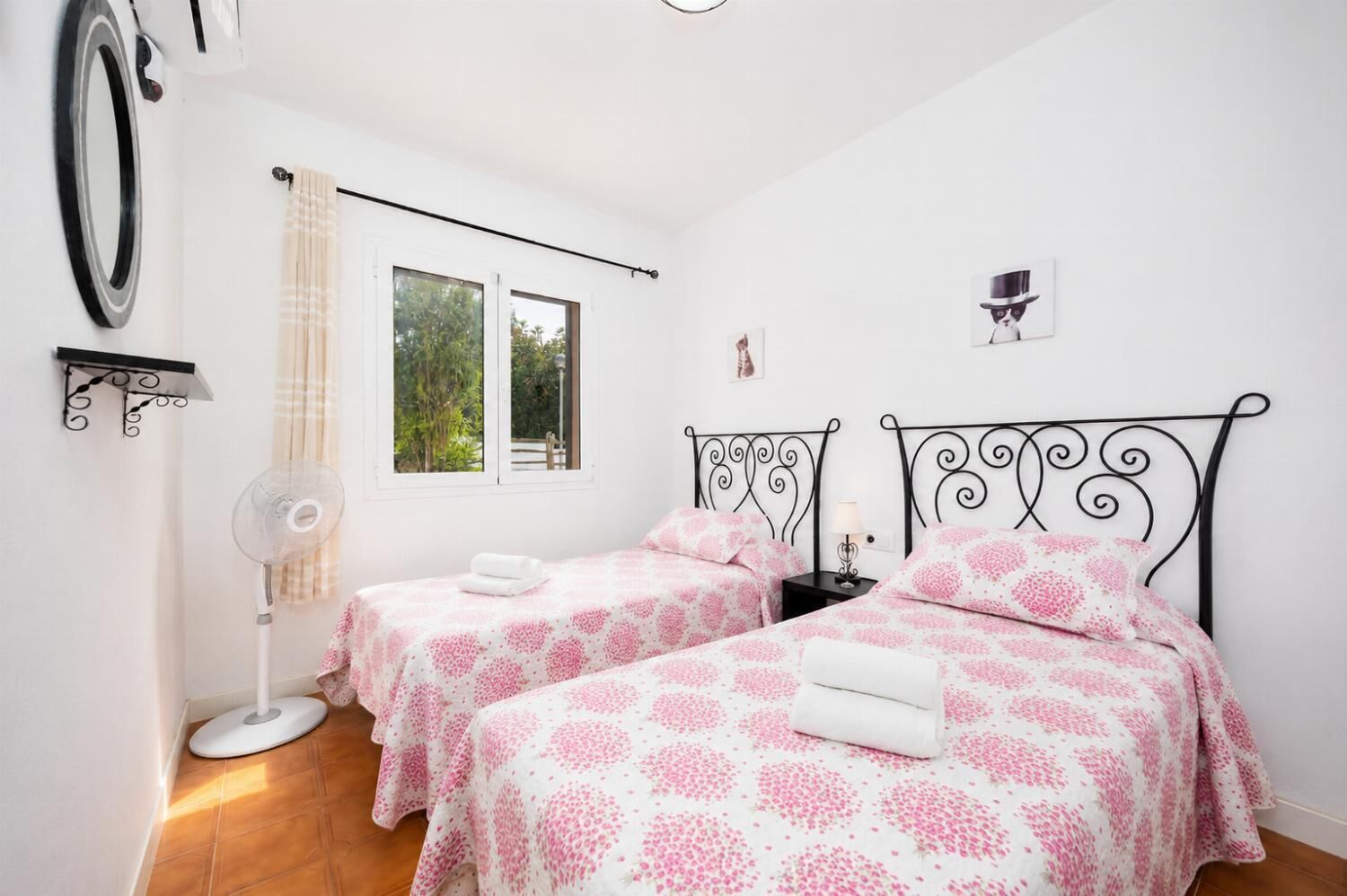 Une chambre avec deux lits, une fenêtre, un ventilateur et une étagère fixée au mur.