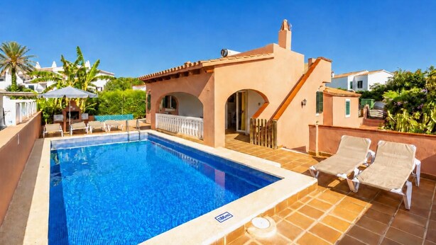 A swimming pool with lounge chairs, a small building with an archway, and palm trees in the background.