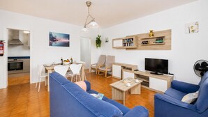 Un soggiorno con divani blu, una televisione, un'area pranzo con sedie, una cucina con forno e una libreria.