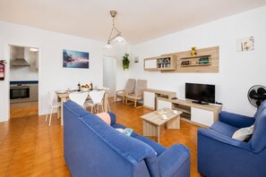 Een woonkamer met blauwe banken, een televisie, een eethoek met stoelen, een keuken met oven en een boekenkast.
