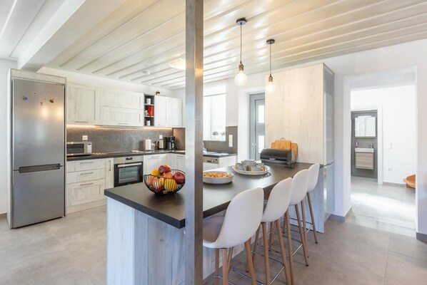 A modern kitchen with white cabinetry, a stainless steel refrigerator, a central island with seating, and pendant lighting. - Villa Constadina Large Private Pool, A/C, WiFi (Paxi)