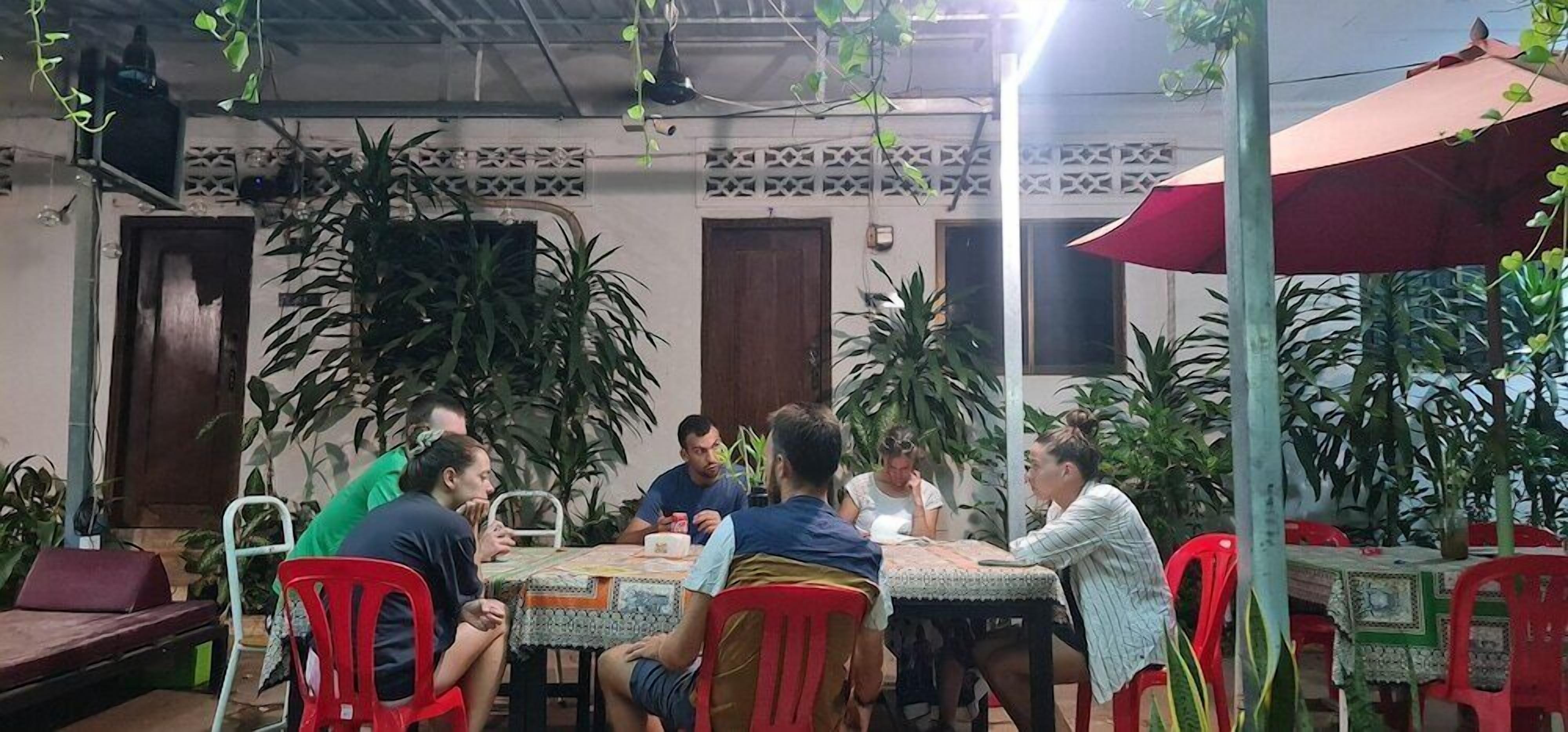 A group of people seated around a table in an outdoor dining area with potted plants and a red umbrella.