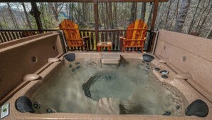 Ein Whirlpool mit zwei Adirondack-Stühlen und einem kleinen Tisch, umgeben von einer Holzterrasse mit Blick auf einen Wald.